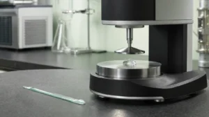 The measuring head of a cone-and-plate rheometer with a bead of clear viscous gel on its polished plate on a laboratory bench