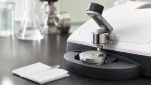 A small drop of clear sample on the ATR crystal of an FTIR spectrometer beneath its pressure clamp, with a glass pipette on a folded wipe beside it
