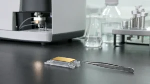 A gold-surfaced biosensor chip in a clear holder beside fine stainless tweezers on a laboratory bench